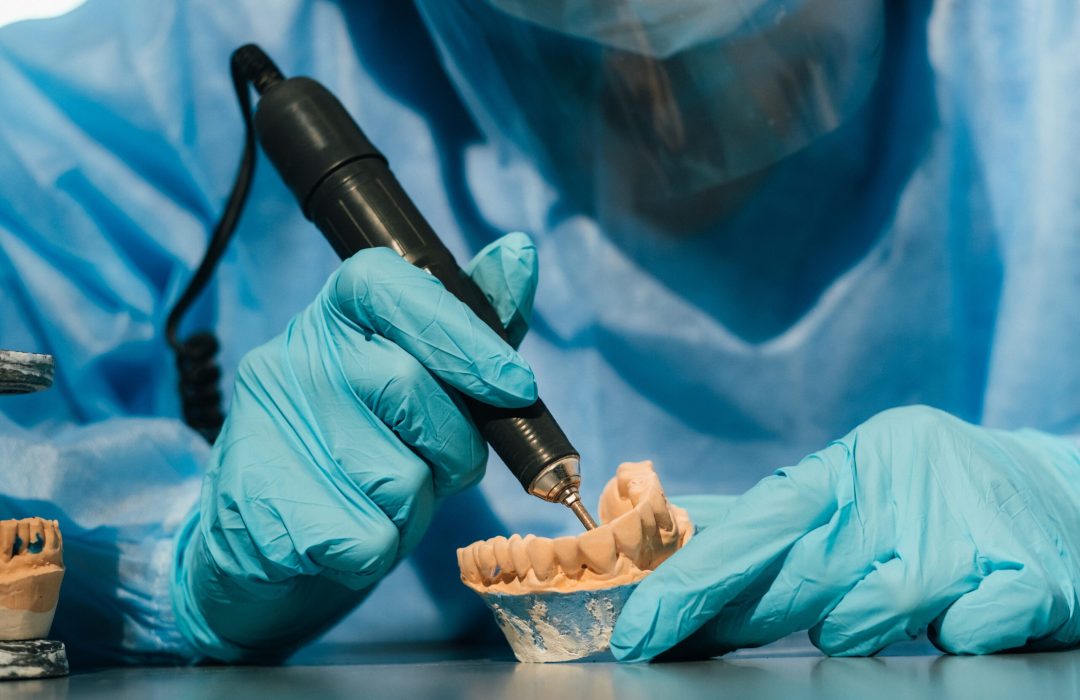 A masked and gloved dental technician works on a prosthetic tooth in his lab.